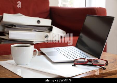 Laptop, Kaffeetasse und Gläser auf einem Wohnzimmertisch und viele Ringordner auf dem Sofa dahinter, die während des Pand in einem obligatorischen Home Office arbeiten Stockfoto