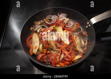 Dampfende Pfanne mit Bratgemüse wie King Auster Pilze, rote Paprika und Zwiebeln auf einem schwarzen Herd, Kopierraum, ausgewählten Fokus, schmal Stockfoto