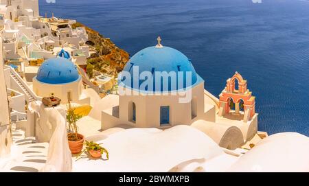 Die wunderschönen und ikonischen blauen Kuppeln von Santorini, einer griechischen Insel, mit Blick auf die Ägäis und den kontrastreichen pfirsichfarbenen Glockenturm. Stockfoto