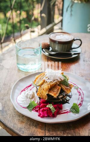 Stapel veganer Pfannkuchen mit Banane, Scheibe Drachenfrucht, Schokolade und Kokosnuss-Creme mit Kaffee und Wasser auf dem Tisch dekoriert Stockfoto