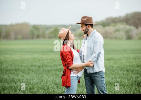 Porträt einer schwangeren Frau mit ihrem Freund, der lässig gekleidet ist und Hüte auf dem Grünfeld zusammen stehen. Glückliches Paar erwartet ein Baby, junge Familie Konzept Stockfoto