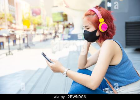 Thai asiatische Mädchen teen allein sitzen für Selbstisolation bei Siam öffentlichen Raum im Freien Musik hören von Smartphone Ware Gesichtsmaske, New normal lifestyl Stockfoto