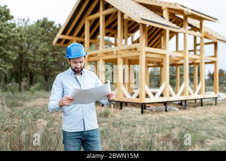 Architekt oder Baumeister stehen mit Blaupausen in der Nähe der Holzhausstruktur auf der Baustelle im Freien. Konzept der Bau Rahmenhäuser aus Holz Stockfoto