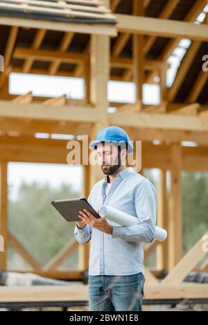 Porträt eines Architekten oder Bauherrn mit digitalem Touchpad vor der Holzhausstruktur. Bau und Gestaltung von Holzrahmenhaus Stockfoto