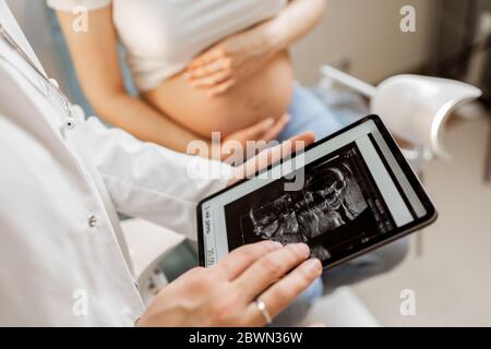 Arzt mit Ultraschall-Scan des ungeborenen Kindes auf einem digitalen Tablet während einer Untersuchung mit einer schwangeren Frau im Büro, beschnittene Ansicht ohne Gesichter Stockfoto