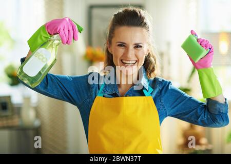 Lächelnde moderne Frau in orange Schürze und rosa Gummihandschuhe mit Spray Flasche von umweltfreundlichen Reinigungsmitteln und grünen Schwamm zeigt Bizeps in der Stockfoto