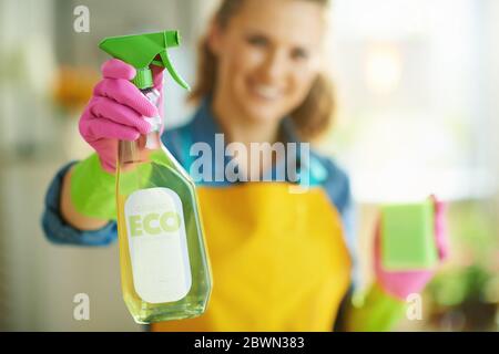 Nahaufnahme auf glückliche Frau in orange Schürze und rosa Gummihandschuhe mit grünen Schwamm zeigt Spray Flasche von umweltfreundlichen Reinigungsmitteln zu Hause in der Sonne Stockfoto