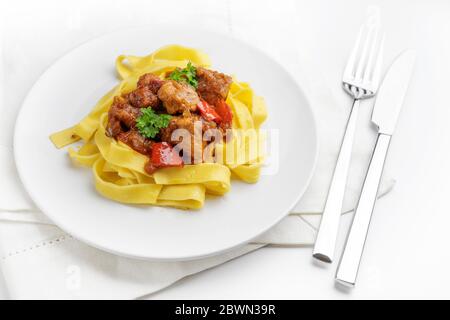 Eintopf oder Gulasch oder aus Schweinefleisch mit Zwiebeln und Paprika auf Tagliatelle Nudeln, Teller, Serviette und Besteck auf einem weißen Tisch serviert, ausgewählte Focu Stockfoto