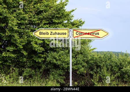 Gelbe Straßenschilder zeigen in entgegengesetzte Richtungen mit deutschem Text Bleib Zuhause bedeutet, und Coronavirus, ländliche Landschaft im Hintergrund Stockfoto