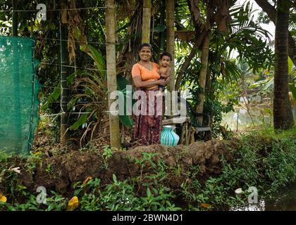 Lächelnd GAP-Zahned Frau hält doe-eyed Baby geschmückt mit Goldohrings und Schmuck und steht am Rande eines Wohnkanals in Keralas Backwaters. Stockfoto