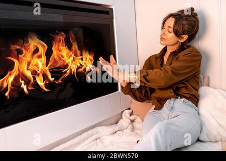 Junge Frau, die sich am Kamin entspannt, die Hände im modernen Wohnzimmer zu Hause wärmt Stockfoto