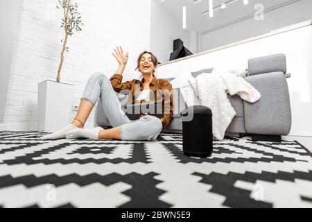 Frau, die Sprachbefehle verwendet, um ein Smart-Home-Gerät zu steuern, das mit einem Smart-Lautsprecher und einem Tablet auf dem Boden im Wohnzimmer sitzt Stockfoto