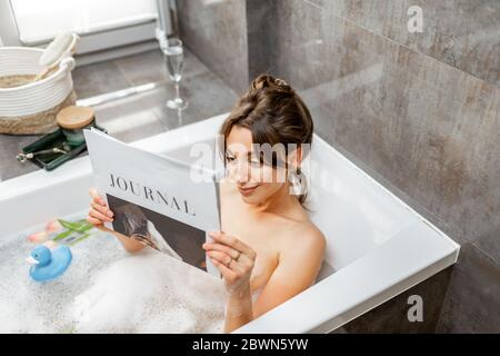 Junge entspannte Frau, die ein Bad nimmt, in der Badewanne liegt und zu Hause ein Magazin liest Stockfoto