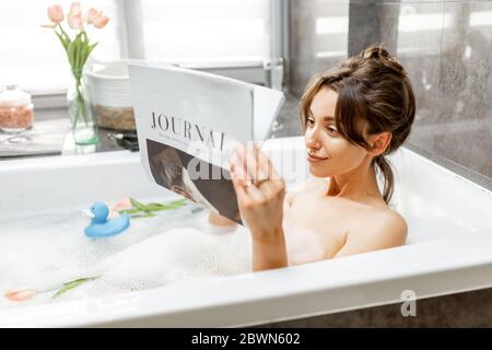 Junge entspannte Frau, die ein Bad nimmt, in der Badewanne liegt und zu Hause ein Magazin liest Stockfoto