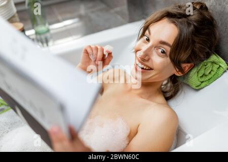 Porträt einer jungen entspannten Frau, die ein Bad nimmt, in der Badewanne liegt und zu Hause ein Magazin liest Stockfoto