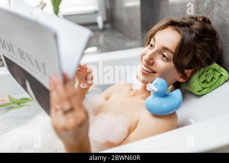 Junge und fröhliche Frau, die sich beim Baden entspannt, mit Gummiente liegt und zuhause ein Magazin liest Stockfoto