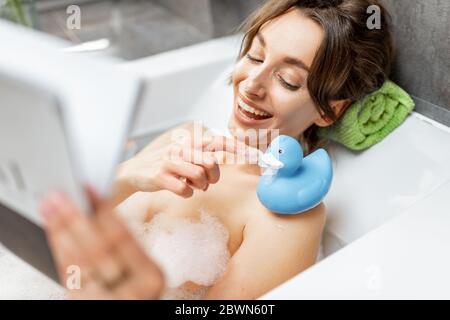 Junge und fröhliche Frau, die sich beim Baden entspannt, mit Gummiente liegt und zuhause ein Magazin liest Stockfoto