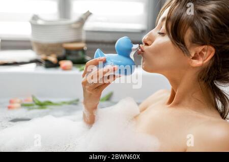 Junge und fröhliche Frau, die ein Bad nimmt, in der Badewanne mit Gummiente liegt, sich im Bad zu Hause entspannt Stockfoto