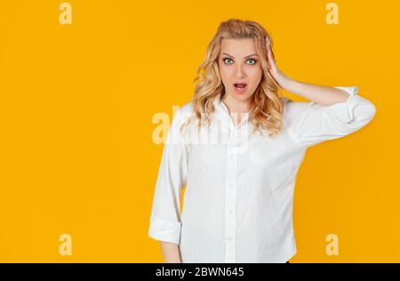 Europäische junge Frau blonde in Panik und Schock verärgert, offenen Mund enttäuscht, berühren ihren Kopf verärgert, schlechte Nachrichten zu hören, stehen verärgert, gelben Hintergrund Stockfoto
