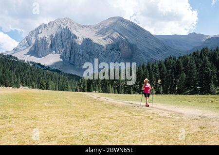 Wandererin mit Strohhut, Shorts und Rucksack auf dem Weg über eine Ebene Wandern mit erstaunlichen Bergen im Hintergrund, während die natura genießt Stockfoto