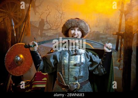 Eine Frau beißt einen Säbel, gekleidet in Kettenpost, Mantel und Pelzhut vor dem Hintergrund von Waffen, einen Zweig mit Krähe und eine Burg in der Ferne Stockfoto