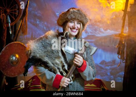 Eine lächelnde Frau, die in Kettenpost und Fell gekleidet ist, mit einer Axt in den Händen vor dem Hintergrund der Arme und Berge in der Ferne Stockfoto