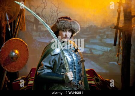 Eine Frau in Kettenpost, Mantel und Pelzhut gekleidet mit einem Säbel in den Händen vor dem Hintergrund von Waffen, einem Zweig mit Krähe und einem Schloss in Stockfoto