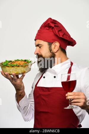 Mann mit Bart isoliert auf weißem Hintergrund. Kochen mit zufrieden Gesicht in Burgund Uniform hält Schüssel gebacken Gericht und ein Glas Wein. Catering und Essen gehen Konzept. Der Koch riecht nach Kartoffelauflauf Stockfoto