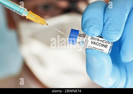 Man Hand in blauen Nitril Handschuh halten kleine Fläschchen mit Coronavirus-Impfstoff, um es mit grün orange Spritze, verschwommenes Gesicht weißen Virusmaske im Rücken injizieren Stockfoto