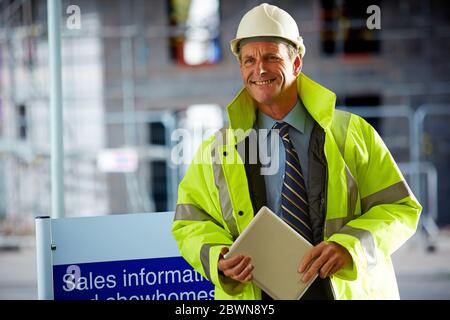 Porträt eines lächelnden Architekten, der einen Hardhat und reflektierende Kleidung trägt, die ein digitales Tablet auf der Baustelle hält Stockfoto