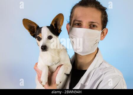 Ein junger Tierarzt in einem weißen Fell und Gesichtsmaske hält einen kleinen Hund oder Welpen, der auf die Kamera schaut Stockfoto