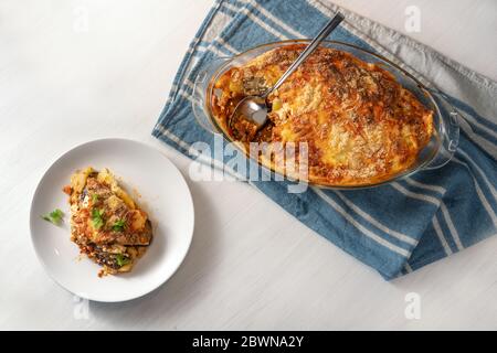 Moussaka, in einer Glasform auf einem blauen Küchentuch gebacken und auf einem weißen Teller serviert, traditionelle griechische Gericht mit Auberginen, Kartoffeln, Hackfleisch Stockfoto