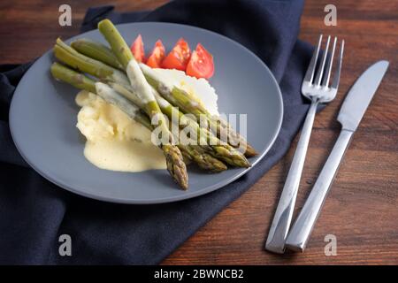 Grüner Spargel auf Kartoffelpüree mit heller Hollandaise-Sauce und Tomaten auf blauem Teller, Serviette und Besteck auf einem dunklen rustikalen Holztisch, auswählen Stockfoto