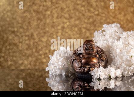 Chinesischer lachender buddha Hotei oder Budai, eingerahmt von weißen Fliederblumen auf goldenem Bokeh-Hintergrund mit Spiegelung Stockfoto