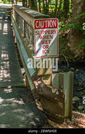 Eine geschützte rostige Metallschiene auf einer Holzbrücke, die über einen Bach führt, mit Warnschildern zur Vorsicht, wenn man nasse oder eisige Nahaufnahme betrachtet Stockfoto