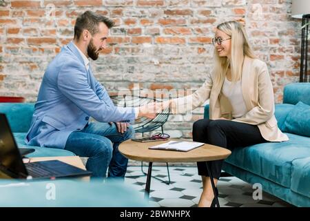 Business, Vereinbarung und Handshake-Konzept. Zwei zufriedene junge männliche und weibliche kaukasische Geschäftspartner, die am Tisch sitzen und sich die Hände schütteln Stockfoto