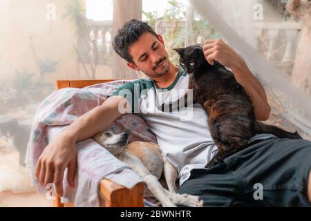 Ein junger Mann, der in einem Sessel im Garten sitzt, streicht einer schwarzen Katze den Kopf Stockfoto