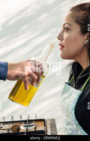Frau trägt eine Schürze riecht hausgemachtes Olivenöl in einer großen Glasflasche, die von der Hand eines Mannes gehalten wird Stockfoto