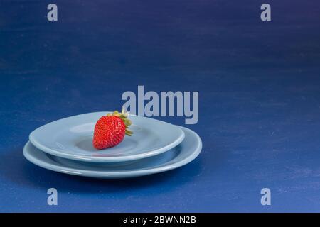 Eine kleine reife rote Erdbeerbeere liegt in einem Teller auf blauem Grund. Kohlenhydratarme Ernährung Stockfoto