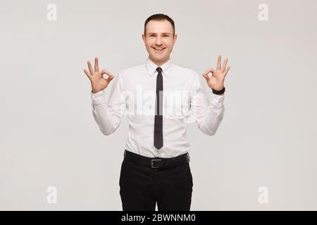 Gutaussehender lächelnder Geschäftsmann in einem weißen Hemd und schwarzer Krawatte zeigt ein OK-Zeichen mit Fingern über weißem Hintergrund, isoliert. Werben Sie für ein Modell Stockfoto