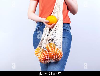 Junge Frau in blauen Jeans und orange T-Shirt ist auf hält orange und String Tasche mit verschiedenen Zitrusfrüchten auf grauem Hintergrund. Konzept Null Abfall, umweltfreundlich Stockfoto