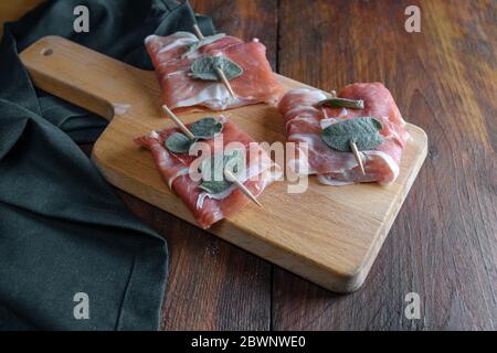 Zubereitetes Hähnchen Saltimbocca mit luftgetrockneten Schinken und Salbeiblättern auf einem Küchenbrett und auf einem rustikalen Holztisch, ausgewählten Fokus eingewickelt Stockfoto