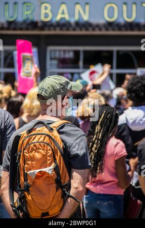 05-30-2020 Tulsa USA - Mann in Mütze mit orangefarbenem Rucksack und Covid-Maske steht hinter der Menge der Protestanten mit Urban Outfitters im Hintergrund Stockfoto
