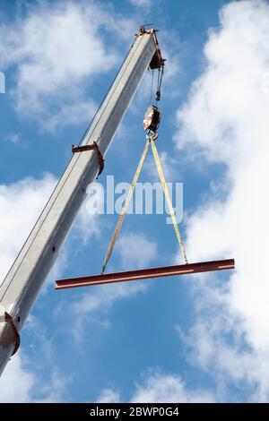 Baukran mit einem Haken und einem Metallprofil, das an Seilen hängt, die hochheben, niedrige Winkelansicht mit wolkenverhangen, sonnigen blauen Himmel Stockfoto