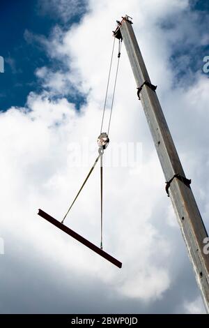 Baukran mit einem Haken und einem Metallprofil, das an Seilen hängt, die hochheben, niedrige Winkelansicht mit wolkenverhangen, sonnigen blauen Himmel Stockfoto