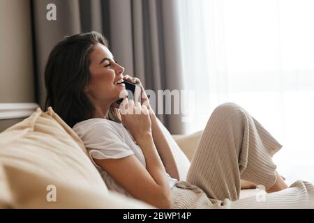 Schöne lächelnde junge Brünette Frau, die sich zu Hause auf einer Couch entspannt, mit dem Handy spricht Stockfoto