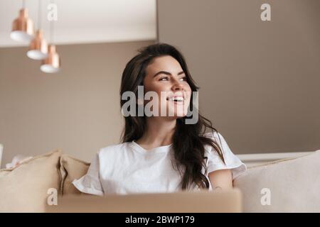 Schöne lächelnde junge Brünette Frau, die sich auf einer Couch zu Hause, mit Laptop-Computer Stockfoto