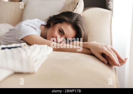 Schöne lächelnde junge Brünette Frau, die sich auf einer Couch zu Hause, Blick auf die Kamera Stockfoto