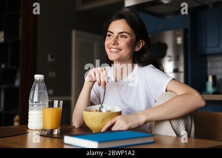 Attraktive lächelnde junge Brünette Frau mit einem gesunden Frühstück, während sie am Küchentisch sitzt Stockfoto