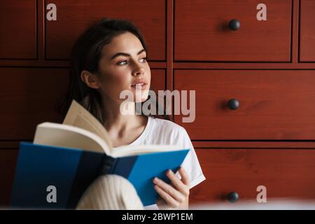 Attraktive lächelnde junge Brünette Frau ein Buch lesen, während sie auf einem Boden sitzen und sich auf einen Schrank lehnen Stockfoto